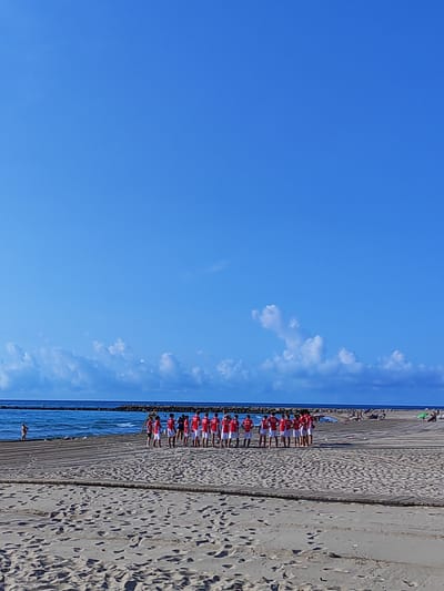 A group of at least 13 young people, likely male, are standing in a line on a wooden boardwalk at the beach in El Campello, Spain. They are all wearing matching red t-shirts with white lettering and white shorts. The boardwalk is set on a sandy beach with the Mediterranean Sea visible in the background. The sea is calm with a breakwater extending into it. The sky is bright blue with scattered white clouds. In the distance, other beachgoers are visible, some relaxing on the sand and others near the water. The overall scene depicts a sunny day at the beach, with the group possibly preparing for or having just finished a sports activity.