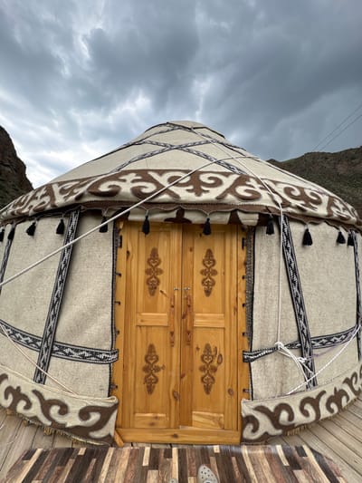 The image shows a traditional yurt, a portable dwelling, set against a backdrop of cloudy skies and mountainous terrain. The yurt is made of felt and fabric, adorned with intricate patterns in brown and white. A wooden door with decorative carvings is centrally located on the yurt's exterior. Ropes are used to secure the yurt, and tassels hang from the upper portion of the structure. The yurt is positioned on a wooden platform, suggesting it is part of a campsite or tourist facility. The scene appears to be in Kyzyl-Korgon, Kyrgyzstan, indicated by the context. No people are visible in the frame. The environment is natural and rugged, consistent with a rural or mountainous region.