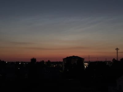 This image captures a twilight scene over a rural area with distant buildings and structures visible. The sky displays a gradient of colors, transitioning from a deep blue at the top to a pale orange and pink near the horizon, indicating the sun has recently set. In the foreground, the silhouettes of trees and dark structures dominate, obscuring details of the immediate environment.
In the middle ground, a large, dark, multi-story building is silhouetted against the brighter sky. To the right of this building, a tall, lattice-like tower, possibly a radio mast or observation tower, stands prominently. In the distance, lights from buildings and streetlamps suggest a settlement. The overall impression is one of a quiet evening in a landscape that blends natural elements with man-made structures. The location is described as Pratasevichi Village Council, Belarus.
