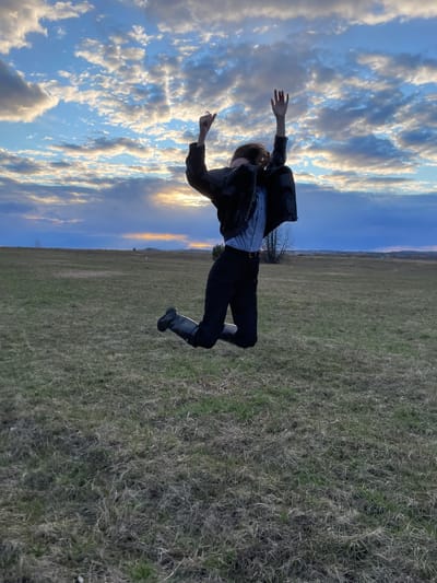 The image, oriented horizontally as if rotated 90 degrees clockwise, captures a person in an open field, seemingly suspended mid-air against a vast, cloudy sky. The individual, dressed in a black jacket, a light-colored collared shirt, dark pants, and dark boots, has their arms outstretched and legs bent, conveying a dynamic, weightless pose. Their hair appears to be light brown or reddish.
The background is dominated by an expansive sky on the left and an uncultivated landscape on the right. The sky is largely blue, filled with scattered white and grey cumulus and stratocumulus clouds. Patches of these clouds are illuminated by the warm, golden light of a setting or rising sun, indicating late afternoon or early evening. The ground, which appears vertically on the right side of the frame, is a large, open field covered with dry, sparse grass, interspersed with greener patches. A faint tree line is visible on the distant horizon.
The scene evokes a sense of freedom, playfulness, or an unusual perspective. No text is visible in the image. This outdoor scene is situated in Kueda, Russia.