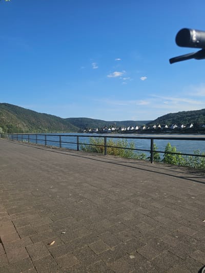 The image depicts a scenic view of the Rhine River in Braubach, Germany, on a clear, sunny day. A paved walkway runs along the foreground, bordered by a black metal railing. Beyond the railing, the wide, blue Rhine River stretches into the distance, reflecting the sky. On the opposite bank, a picturesque village with white-roofed houses is nestled at the base of lush, green, tree-covered hills. The sky is a vibrant, cloudless blue, with a few wispy clouds visible. In the upper right corner of the image, a blurry, black object, likely part of a camera or bicycle handlebar, is visible. The overall impression is one of tranquility and natural beauty.
