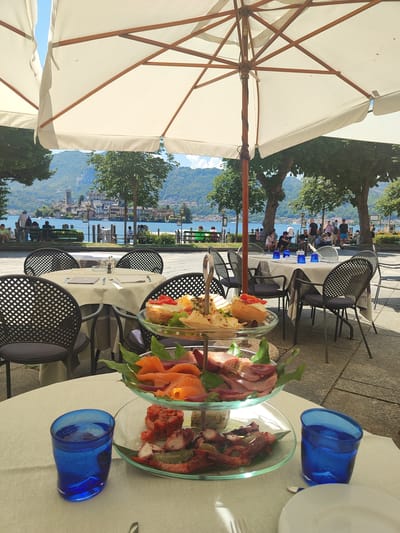 The image captures an outdoor seating area of a restaurant in Orta San Giulio, Italy, with a picturesque view of the lake and the island of San Giulio in the background. The foreground features a table set with a white tablecloth, two blue glasses filled with water, and a three-tiered platter of assorted food, including smoked salmon, thinly sliced meats, and seafood like octopus and shrimp. In the middle ground, several tables with chairs are arranged, some covered with white tablecloths and set with cutlery, glasses, and salt and pepper shakers. Beyond the tables, a paved walkway is visible, with trees lining the path and a view of people strolling by the lake. The background showcases the tranquil lake, with boats dotting the water, and the charming town of Orta San Giulio situated on the hillside, culminating in the distinct silhouette of the Basilica of San Giulio on the island. The scene is shaded by large, off-white umbrellas, indicating a sunny day.