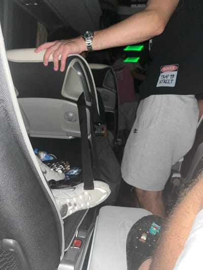 The image shows an interior view of a passenger aircraft. A person is seated in a business class seat, wearing grey sweatpants and a black t-shirt with a "TRAP 19 STREET" graphic. Another person is wearing black Crocs with charms. A third person is wearing a silver watch on their left wrist. An additional pair of Nike sneakers is visible in the storage area near the passenger's feet. The interior is arranged in a semi-private pod layout, common in some premium airline cabins. The location is not identifiable as being in Slavovitsa, Bulgaria, based on the visual information provided.
