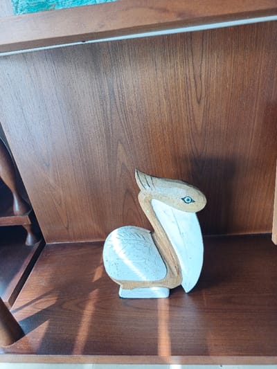 A wooden pelican figurine sits on a dark brown shelf in front of a brown paneled wall. The pelican is mostly beige and white with a light blue eye. The carving has wing details. Part of a wooden shelf unit is visible to the left. The light casts shadows across the shelf.