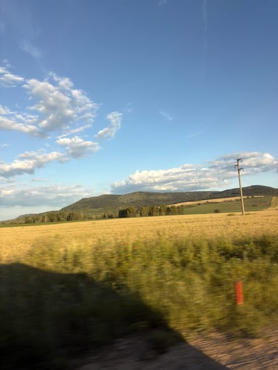 An outdoor, horizontal, eye-level shot depicts a rural landscape under a blue sky with scattered clouds. The foreground is blurred due to motion, showing a shadowy mass on the left and a dirt road on the right. A red marker is visible in the middle ground on the right. The mid-ground features a vast field of dry, golden crops, possibly wheat or barley, with a few red poppy flowers scattered throughout. A line of trees forms a boundary between the field and the rolling hills in the background. The hills are covered with dense vegetation and appear green and brown. A single, tall, gray utility pole stands in the middle of the field on the right side of the image. The overall scene suggests a warm, sunny day in the Bulgarian countryside near Komarevo.