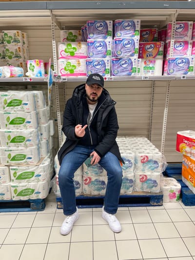 An indoor shot shows a man in a black jacket, blue jeans, and white sneakers sitting on a pallet loaded with packages of "Ultra Moelleux" toilet paper. He's in a supermarket aisle flanked by shelves stocked with various brands of toilet paper like "Lotus" and "Le Trèfle". He is holding a phone and gesturing with his middle finger raised. The floor is tiled. Overhead lighting illuminates the scene. A package of "Sopalir" toilet paper sits on top of the pallet.
