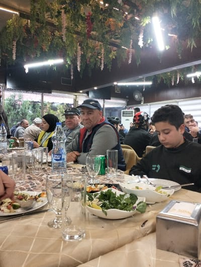 A group of people are gathered around a table filled with food and drinks in what appears to be a restaurant or banquet hall. The table is covered with a tablecloth and various dishes of salad and other foods. Glasses and water bottles are scattered around. The decor includes hanging plants and flowers. The atmosphere suggests a social gathering or a meal being shared by friends or family.
