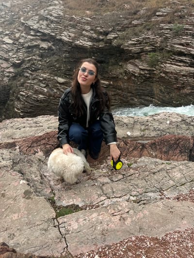 An outdoor scene in a rocky, riverine environment near Mazići, Montenegro, depicts a female individual with a dog. The female, possessing long dark hair and wearing light blue tinted sunglasses, a black leather jacket over a light-colored top, and dark blue jeans, is crouched on reddish-grey stratified rock. Her left hand rests on the back of a small, cream-colored, fluffy dog, which is positioned on the ground in front of her, appearing to sniff downwards. Her right hand holds a yellow and black retractable dog leash connected to the dog. The female looks towards the camera with a "duck face" expression. Behind the subjects, a fast-flowing river with white rapids is visible to the right. The background consists of a steep, layered rock formation or cliff face, indicative of a canyon or gorge. The overall lighting suggests an overcast sky.