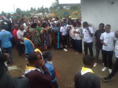 A diverse crowd gathers outdoors. Some wear t-shirts with a circular graphic. Others sport yellow neckerchiefs. Many individuals are dressed casually in everyday attire. In the background, a yellow bus is visible. The setting appears to be a rural area with trees and buildings nearby. The group stands in a loose formation, seemingly waiting or observing something out of frame.
