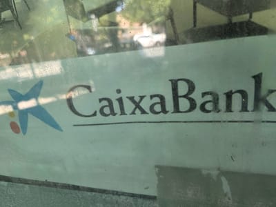 A photograph captures a close-up of a sign with the text "CaixaBank" printed in black, bold, serif font. The sign has a light green background with a slightly faded and textured appearance, possibly due to being on a window or an outdoor surface. To the left of the text is the CaixaBank logo, which features a stylized blue asterisk-like shape with a red dot to its left. The sign is mounted on a surface that reflects the environment, showing blurry shapes of trees, foliage, and possibly buildings. The lower portion of the image reveals a reflective surface, likely glass, with a horizontal line running across it, indicating a frame or shelf. The overall lighting suggests daylight. This sign is located on Calle Real, number 74, in Collado Villalba, Spain.