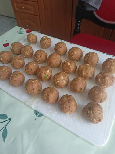 The image shows a white cutting board covered in plastic wrap, with numerous round, light brown meatballs or dough balls arranged on it. The meatballs appear to be uncooked and contain visible specks of nuts or spices, possibly pistachios or chopped nuts. They are arranged in rows, filling most of the surface of the cutting board.
The cutting board is placed on a table covered with a light green tablecloth that has a pattern of dark green leaves and stems. In the background, a wooden kitchen cabinet with drawers and handles is visible, suggesting an indoor setting, likely a kitchen. To the right of the cabinet, part of a dark chair with a red seat is visible. The lighting appears to be natural, suggesting daytime. There is no visible text in the image. The overall impression is that of food preparation.