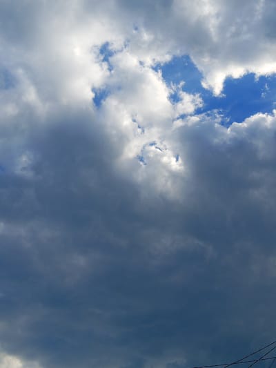 The image captures a dramatic sky filled with a mix of dark, heavy storm clouds and patches of bright blue sky. The clouds are varied in texture and color, ranging from deep grays and blues to lighter whites where sunlight attempts to break through. Some clouds appear thick and ominous, while others are thinner and wispy.
The scene is an outdoor, natural setting, focusing solely on the sky. There are no visible people or man-made objects in the immediate foreground, except for a hint of thin, dark wires in the bottom right corner, suggesting the presence of infrastructure. The lighting indicates it is daytime, likely late afternoon or early evening, given the contrast between the bright sky and the shadowed clouds, which could suggest an approaching storm or the aftermath of one.
The overall mood conveyed by the sky is one of dynamic weather and potential change, evoking a sense of awe or perhaps foreboding due to the intensity of the cloud formations. The location context of Panino, Russia, places this dramatic sky in a specific geographical setting, though no explicit visual cues within the image pinpoint this location beyond the provided context. There is no visible text within the image.