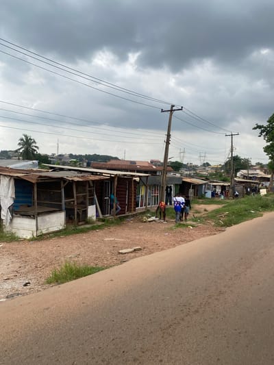 This image depicts a street scene in Sogeke, Nigeria, under a cloudy sky. The perspective is tilted, with the ground appearing on the right and the sky on the left. Along the side of a dirt path, which merges into a paved road on the right, are several structures that appear to be shops or dwellings. These buildings are a mix of corrugated metal and wood, some with open fronts revealing interior spaces.

Several people are visible, mostly standing or walking along the path. In the foreground, a group of young men are gathered, one wearing a blue and white jersey. Further down the path, other individuals are seen near the shops. One person is sitting in what looks like a doorway of one of the buildings.

Power lines crisscross the scene, supported by wooden utility poles, indicating the presence of electricity infrastructure. In the background, a cluster of buildings can be seen on a hill, with more power lines and transmission towers visible. A palm tree is also present in the mid-ground.

The weather appears overcast and possibly humid, consistent with a tropical climate. The overall impression is of a busy, informal marketplace or residential area, with daily life unfolding along the roadside. No specific text is legible in the image.