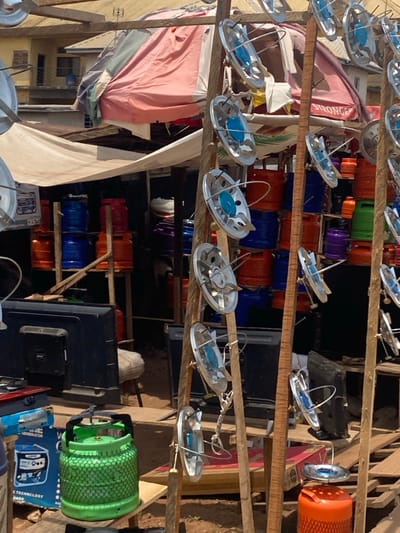 The image depicts an outdoor market stall in Afikpo, Nigeria, under bright daylight. In the foreground, a green gas cylinder with a black handle and a protective mesh is centrally positioned on a wooden surface. To its left, a blue box labeled "SYNERGY GENERATOR" supports a small, silver-colored single-burner gas stove. On the right, two vertical wooden poles are used as display stands, each exhibiting multiple small, round, silver-colored gas stove burners with blue central components attached by wires. A partial view of a person's torso and arm is visible on the far left. At the bottom right, an orange gas cylinder is topped with another small burner.
In the midground, behind the immediate display, several black flat-screen televisions are stacked. Further back, under two fabric awnings – one light-colored and another reddish-pink – numerous gas cylinders of varying colors (orange, blue, purple, and green) are vertically stacked on wooden shelves. The ground is exposed reddish-brown earth. In the distant background, a multi-story building with a light-colored facade and a brown roof is visible under a clear sky. The scene indicates a retail environment specializing in gas cooking equipment and related appliances.
