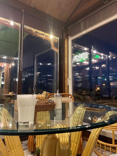 The image displays an indoor scene within a cafe or restaurant in Yantarny, Russia, observed during nighttime. The foreground features a clear glass table with a subtle greenish edge. On the table, from left to right, are a tall glass containing a milky white beverage with a black straw, a small brown woven basket, and a silver-topped dispenser filled with white granules. A dark-cased smartphone rests on the far right of the table surface.

The interior environment includes reflections of golden, possibly wicker or rattan, chairs that serve as seating. The ceiling features visible wooden beams, and warm artificial lights are present, some appearing directly as bright bulbs affixed to a brown pipe-like structure running along the upper wall. A long, thin, metallic chain or pull cord hangs vertically near a window pane on the left.

The background consists of large windows overlooking a dark exterior, indicating night. Through the glass, distant city or settlement lights are visible. A green neon sign, appearing as the reversed characters "TGNIP" in reflection, illuminates a section of the window. No individuals are visible in the scene.