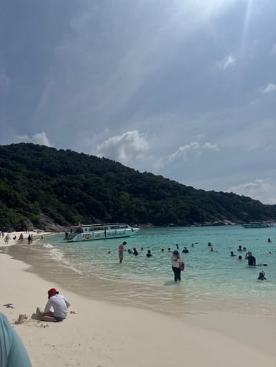 The image depicts a beach scene in Lam Kaen, Thailand. A lush, green hill rises along the coastline. The turquoise water is filled with numerous people swimming and wading. A boat labeled "LOVE Andaman" is docked at the edge of the water. The beach has light, fine sand. On the sand, a person wearing a white shirt and red cap is digging. The sky is partially cloudy with a light blue hue.