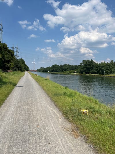 This is a vertical, eye-level, outdoor, daytime photograph of a gravel path alongside a canal in Herne, Germany. The path is in the foreground and extends into the distance. To the right of the path is a body of water, the canal, with rippling, dark water. The far bank of the canal is lined with trees. In the background, there is a wind turbine with red and white blades, and a red bridge behind it. Tall electricity pylons are visible on the left side of the image, also to the left of the trees. The sky is blue with scattered white clouds. There are a few people visible in the distance on the path and in the canal. Two individuals are lying on the grass next to the path on the canal's edge.