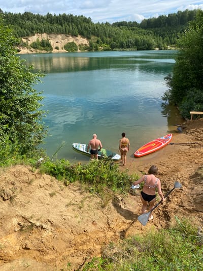A group of four people are engaged in recreational activities by a lake. Two men and two women are present. One man, partially submerged in the water, stands beside a paddleboard. A woman in a yellow bikini stands on the sandy shore, near a bright orange paddleboard. Another woman, wearing a black and white bikini, ascends a sandy embankment, carrying two paddleboard paddles. The scene is set in a natural environment characterized by a clear blue lake, surrounded by lush green trees and sandy cliffs. A wooden bench is visible in the background. The image appears to be taken on a sunny day, with clouds in the sky. The location is identified as Plintovka, Russia.