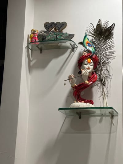 This image displays a section of a white interior wall in a residence or establishment located in Surat, India. Two clear glass shelves, supported by silver-colored metal brackets, are mounted on the wall.
The upper shelf holds a collection of decorative items: On the left, a pair of small, brightly colored figurines depict a male and female couple in traditional attire, with the female figure in a pink dress and the male in a yellow garment. Behind them, a green-tinted decorative piece, possibly resin, resembles a landscape with trees. To the right, two rustic, heart-shaped plaques are visible, one inscribed "SANGITA" and the other "SITU".
The lower shelf features a statue of Lord Krishna, rendered in white with prominent red and gold accents on his attire and turban. A vibrant, multi-colored peacock feather is integrated into his turban. The statue depicts Krishna playing a flute. To the right of the statue, a collection of natural, dark peacock feathers is arranged vertically against the wall. Shadows from the shelves and objects are cast onto the wall, suggesting illumination from an external or overhead light source.