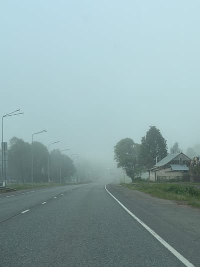 A road scene shrouded in heavy fog. A gray asphalt road with a white dividing line runs along the right side of the frame. Streetlights line the left side of the road, disappearing into the fog. Trees are visible alongside the road and behind the streetlights, their details obscured by the fog. A building, partially hidden by the trees and fog, is visible near the top right of the image. The pervasive fog reduces visibility to a minimum. The location is indicated as Staraya Yum'ya, Russia.