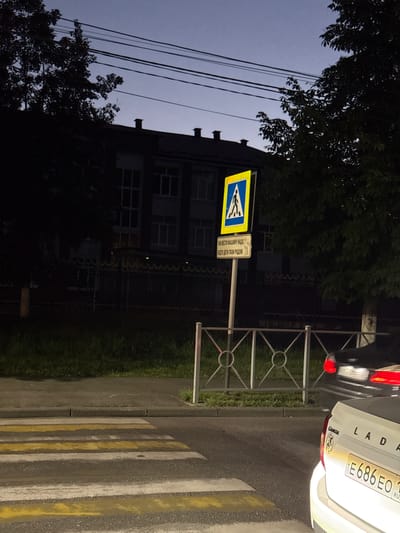 This media file captures an outdoor scene at dusk, likely in Charavali, Russia. The primary subjects are a pedestrian crossing with its distinctive yellow and white striped markings, a street sign indicating a pedestrian crossing and a supplementary sign with Russian text, a section of a building in the background, and a car with a Russian license plate. The car, a Lada, is partially visible, showing its rear end and license plate "E686EO 1". A metal barrier, possibly part of a fence or railing, is positioned near the pedestrian crossing and sign. Lush green grass lines the side of the road. Overhead, a tangle of power lines stretches across the frame, silhouetted against a twilight sky. The building in the background appears to be a multi-story structure with visible windows. The lighting suggests it is either early morning or evening, with artificial lights from the car and potentially streetlights illuminating the scene. The overall mood is calm and ordinary, depicting a common urban or suburban streetscape. The Russian text on the supplementary sign reads "ТАК ВЕСТИ МАШИНУ НАДО, БУДЬТЕ ДЕТИ ТВОИ РЯДОМ" which translates to "THIS IS HOW YOU SHOULD DRIVE, YOUR CHILDREN ARE NEARBY."