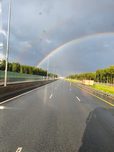 A wet highway stretches into the distance, flanked by a line of trees on the right and a sound barrier on the left. A double rainbow arcs across the cloudy sky. Streetlights line the highway. A small, red building is visible on the right side of the road. The highway appears to be in Krutitsy, Russia.