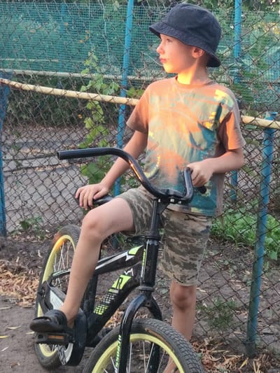 A young boy, wearing a dark bucket hat and a t-shirt with an orange and teal graphic, stands with his foot on the pedal of a black BMX bicycle. He is wearing camouflage shorts and black sandals. The boy is looking off to the right, his face illuminated by the warm, golden light of the setting sun, suggesting it is late afternoon or early evening.
The setting appears to be outdoors, likely a yard or a park area, with a chain-link fence in the background. The fence has a blue metal post and a weathered wooden or metal railing running horizontally. Green foliage and dry leaves are visible around the base of the fence and on the ground. The light filtering through the fence creates dappled patterns on the ground and the boy's clothing.
Visible text on the bicycle includes "CROSSRIDE" and "R" logo. There is also some text on the boy's t-shirt, but it is mostly obscured and illegible. The overall mood is one of a casual, perhaps slightly pensive moment, captured in the soft glow of the golden hour. The location context is Saksahan, Ukraine.