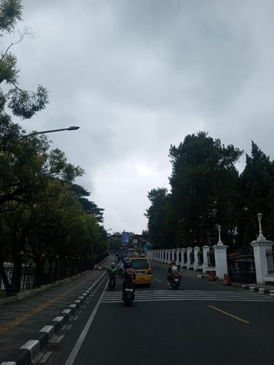 A road under a cloudy sky features a mix of vehicles, including cars and scooters. The road is lined with trees on the left and a white fence with decorative lamps on the right. A sidewalk is visible along the left side of the road, bordered by a black and white curb. Yellow lines mark the road's center. The scene suggests an urban setting, possibly a city street on a slightly overcast day.