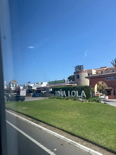 The image depicts a sunny outdoor scene with a clear blue sky and a well-maintained lawn in the foreground. A large white sign on the lawn reads "DOÑA LOLA." Behind the lawn is a row of buildings with white facades and red-tiled roofs, some featuring awnings and signage for various establishments. One prominent building is a round stone tower topped with a sign displaying images of food. To the right of the tower, a building with an arched entrance has the name "Doña Lola" written above it. Cars are parked on a paved area in front of the buildings, and a dark-colored car is visible on the left side of the frame. The scene appears to be a commercial area or a tourist destination. Based on the signage, the location is Doña Lola in Mijas, Spain.