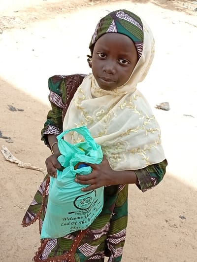 A young girl stands outdoors, holding a teal plastic bag. She wears a patterned green and brown dress with lace trim, and a white hijab adorned with gold embroidery. Her head is covered with a green and white patterned headscarf. The bag in her hands features the words "Welcome You! End Of The Year Class..." along with a logo. The background consists of a sandy terrain.
