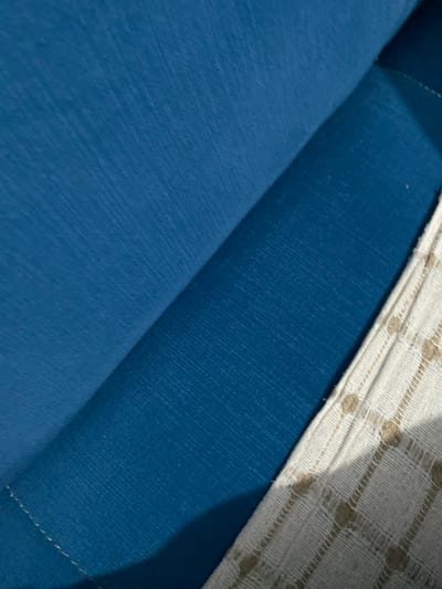The scene, located in Sredkovets, Bulgaria, depicts a close-up view of two distinct textile materials. A section of blue fabric, exhibiting a fine, parallel linear texture, occupies the majority of the frame, spanning the upper-left and central areas. This blue material is folded, creating a prominent diagonal crease from the upper-right to the lower-left, with a subtle shadow cast along the fold. To the right, an off-white fabric is visible, featuring a repeating grid pattern composed of small, raised beige dots or squares. The visible edge of this lighter fabric displays consistent stitching. No individuals or actions are present in the image.