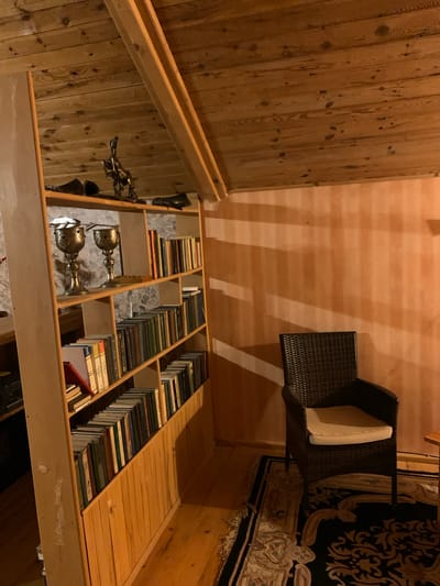 The image depicts an interior scene, likely a room with a sloped wooden ceiling and wooden walls. On the left, there is a multi-tiered wooden bookshelf filled with books of various sizes and colors. Two ornate metal chalices are prominently displayed on one of the shelves, along with a decorative metallic sculpture of a horse and rider above them. To the right of the bookshelf, a dark-colored wicker chair with a light-colored cushion sits on a rug with an intricate pattern. The walls appear to be a warm, peachy-orange color, with some textured vertical lines and shadows cast by an unseen light source. The overall ambiance suggests a cozy, possibly study or reading area. The location in Mepiskalaki, Georgia, is not visually discernible from the provided image.