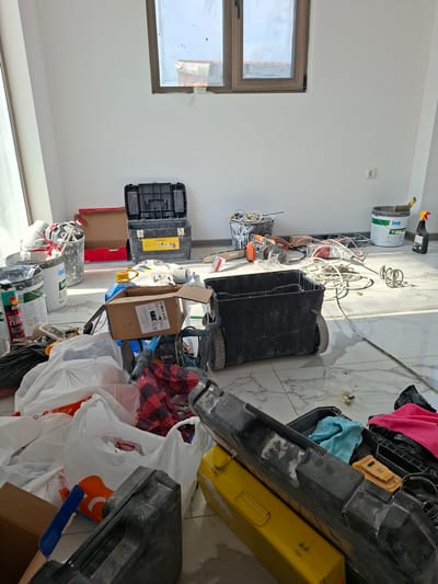 The image depicts an interior scene of a room, likely undergoing construction or renovation, located in Dolna Malina, Bulgaria. The room is sparsely furnished, with white walls and a window with a wooden frame at the top center. The floor is tiled with a white marble pattern.
The floor is covered with various construction materials and tools. Prominently displayed are several toolboxes, some open and containing items, others closed. There are multiple buckets, some containing what appears to be construction mix or residue, and others filled with various tools and debris. Bags, some clear plastic and others with contents like red and black checkered fabric, are scattered across the floor.
Electrical cords are visible, snaking across the floor and connected to tools. Among the identifiable tools are what appear to be mixers, a power drill, and possibly a saw. A large white bucket with a blue label that reads "KNAUF" and "SUPER..." is positioned towards the right side of the image. Next to it is a black spray bottle. A yellow toolbox is visible in the foreground, positioned next to a large black toolbox.
The overall impression is one of an active construction site, with materials and equipment left in disarray, suggesting a pause in work rather than abandonment. No individuals are present in the image. The lighting suggests daytime.