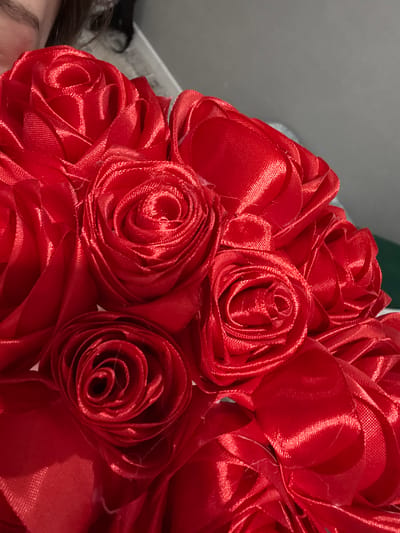 A close-up, eye-level shot showcases a bouquet of vibrant red satin roses. The roses are intricately crafted, with layered petals forming a realistic rose shape. The satin fabric reflects light, creating highlights and shadows that emphasize the texture and depth of the petals. Some loose threads are visible, suggesting the handcrafted nature of the bouquet. In the upper left corner, a sliver of a person's eye, eyebrow, and hairline is visible, indicating the bouquet is being held or presented. The background is a soft blur of gray and a hint of dark green, providing no specific environmental context. The location in Zaşağan, Kazakhstan, is not discernible from the visual information provided.
