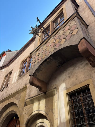 This is a low-angle, exterior shot of an old building in Würzburg, Germany. The facade is made of weathered, light-colored stone and plaster, with several arched entrances visible at the bottom. Above the entrances, there are windows with wooden frames and intricate metal grilles. A distinctive feature is an ornate, diamond-patterned balcony that protrudes from the upper story. Hanging from a metal pole extended from the building is a star-shaped, metallic lantern. The sky above is clear and blue. The architectural style suggests a historical European setting, likely in a town center or older district.