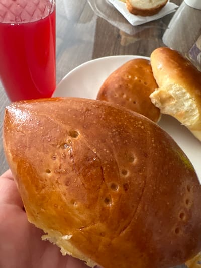 A close-up, eye-level shot shows a hand holding a golden-brown, baked pastry that appears to have small air pockets or dimples on its surface. In the background, on a white plate, are two more similar pastries, one of which is partially broken. To the left, a clear glass filled with a vibrant red liquid is visible. The setting appears to be a table with a reflective surface, suggestive of a home or cafe environment. The image does not provide any information about the location in Znamya Oktyabrya, Russia.