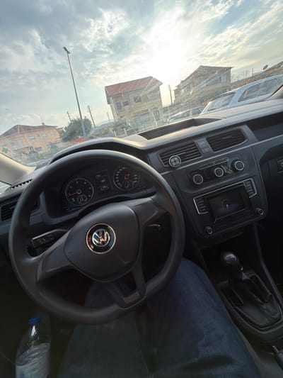 The image depicts an interior-exterior view from the driver's seat of a Volkswagen vehicle, likely a Caddy, located in Felgueiras, Portugal.
**Interior:** The foreground is dominated by the black three-spoke Volkswagen steering wheel. Behind it, the instrument cluster displays a digital time of 13:31 and an ambient temperature of 19.5°C. Analog gauges for engine RPM, speed, and fuel level are visible. The dashboard is dark grey, featuring central air vents, one of which has a small circular, three-pronged accessory clipped into it. Below the vents are controls for the climate system and audio, and a central infotainment screen that appears to be active or loading. To the right, a manual gear shifter is visible. The lower left shows a clear plastic water bottle with a blue cap resting on the floor or seat. The driver's legs, clad in blue denim jeans, are visible below the steering wheel and extending to the right.
**Exterior:** Through the windshield, the scene is illuminated by strong sunlight from the upper right, resulting in significant lens flare and glare. The sky is partly cloudy with patches of bright blue. In the mid-ground, a two-story building with a reddish-brown tiled roof and light-colored, possibly worn, facades stands prominent, slightly right of center. To its left, another multi-story building with a light beige or cream facade and a tiled roof is partially visible. A tall, dark street light pole is positioned to the left of the central building. In the background, parked vehicles and additional structures are discernible, indicating an urban or semi-urban environment. Utility poles and lines are also present in the distance. The road surface or parking area appears to be asphalt.