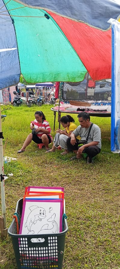 In an outdoor setting in Bandasari, Indonesia, three adults are observed resting under a multi-colored tarp providing shade. The ground beneath them is covered in green grass. To the left, an adult female sits on a small dark stool, wearing a red and white striped shirt and dark pants, and is looking down at an object in her hands. Beside her, another adult female sits on the grass, dressed in a yellow shirt and light-colored pants, focused on a smartphone with an earbud in her right ear. To her right, an adult male crouches, wearing a grey T-shirt with a dark symbol, dark pants, and sandals, holding a lit cigarette and looking downwards. In the foreground, a dark grey plastic laundry basket contains multiple coloring pages with colorful borders, the top page displaying an outline drawing of two smiling dolphins with bubbles. In the background, beyond the resting individuals, other market stalls with merchandise and several parked motorcycles are visible.