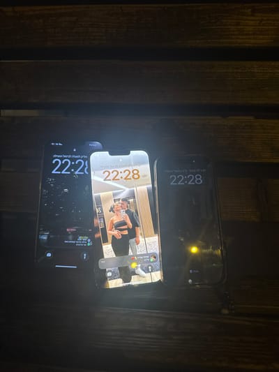 The image depicts an indoor scene within the city of Azor, Israel. Three smartphones are displayed horizontally on a dark, horizontally slatted wooden surface. The ambient lighting is dim, with light primarily emanating from the phone screens.
From left to right:
1. The leftmost smartphone displays a lock screen with the time "22:28" in white text, accompanied by Hebrew text above and below. A "5G" network indicator is visible. The background image is dark and indistinct, showing faint reflections.
2. The central smartphone, brightly illuminated, displays a lock screen with the time "22:28" in orange text. Its wallpaper is a mirror selfie showing a female in a black two-piece outfit holding a phone, standing alongside a male in a dark top and light pants. They are positioned in an area with vertical light wood paneling, likely an elevator or mirrored room. A Hebrew notification bubble is present at the bottom of the screen. A "5G" network indicator is visible.
3. The rightmost smartphone displays a lock screen with the time "22:28" in white text, with Hebrew text above. A "5G" network indicator is visible. The background is predominantly dark, featuring a circular yellow reflection near the bottom center.
All three phones display the identical time of "22:28". No direct human presence or interaction is observed within the immediate environment of the phones; the only people depicted are within the wallpaper image on the central phone.