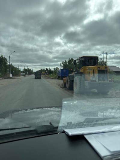 The media file depicts a scene on a road in Parma, Russia, likely during daytime due to the visible daylight. The sky is overcast with heavy, gray clouds, suggesting a cloudy or potentially rainy day. The road appears to be under construction or recently paved, with visible dirt and debris along the sides.

The primary objects in the foreground are construction vehicles. A large, yellow loader, possibly a CAT, is parked on the side of the road. Its license plate is partially visible and reads "2097 MA 11". Next to it is a blue container or truck. Further down the road, other vehicles, including a dark-colored van and possibly trucks, are visible.

The setting appears to be a semi-rural or industrial area with trees lining the road and a building in the background. Streetlights are also visible along the road. The image is captured from inside a vehicle, with a reflection on the window obscuring some details. There are no people clearly visible in the frame. The overall mood is utilitarian and perhaps a bit somber due to the cloudy weather and construction activity.