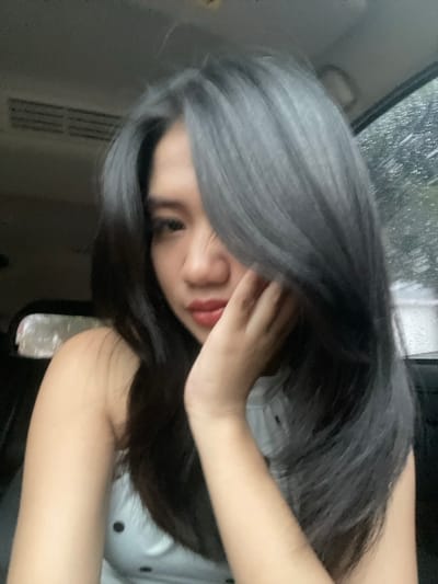 The scene is located within a vehicle in Bojongherang, Indonesia. A young female individual is depicted in a close-up, selfie-style photograph. She possesses long, dark, layered hair, with a prominent section on the viewer's right exhibiting a distinct gray tone, which partially covers her left eye and cheek. Her right eye (viewer's left) is dark and open, and her lips are reddish-pink and slightly parted. Her left hand is raised, with fingers resting against her chin and lower cheek. She is dressed in a light-colored, sleeveless top featuring black polka dots, visible on her left shoulder and chest. The background reveals the interior of a vehicle, including a light-colored ceiling and a dark rectangular air conditioning vent positioned on the upper left. A window to the subject's right displays visible streaks of water, indicating ongoing precipitation outside. The external view through the window is blurred and indistinct.
