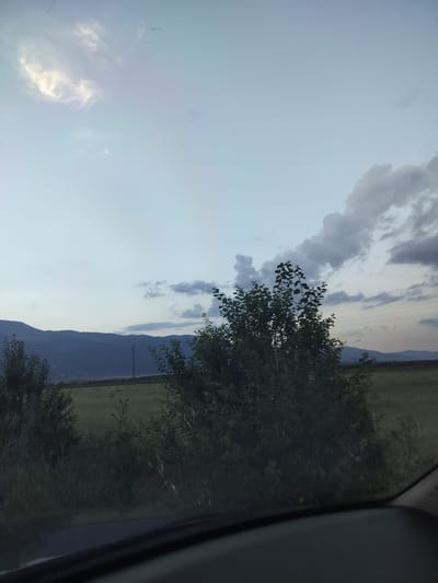 The image depicts a rural landscape scene, likely in or around Manolovo, Bulgaria, captured from within a vehicle. The foreground shows the dark interior of a car, specifically the dashboard and windshield. Beyond the vehicle, a field stretches towards a range of dark, silhouetted mountains in the distance. The sky is a soft gradient of pale blue with scattered clouds, some appearing bright and illuminated, suggesting either early morning or late evening light. To the right and center of the frame, lush green trees and bushes are prominent, partially obscuring the field and mountains. A lone utility pole with wires is visible in the middle ground, to the left of the central trees. The overall atmosphere is serene and natural, characteristic of a countryside setting.