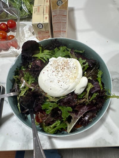 The image depicts a prepared salad in a light blue bowl, positioned on a white countertop, likely within a domestic setting in Cidade da Maia, Portugal. The salad consists of mixed green and red leaf lettuce, several halved cherry tomatoes, and a large, white burrata cheese ball prominently placed in the center, seasoned with red pepper flakes and black pepper. Two silver-colored utensils, appearing to be spoons or forks, rest within the salad bowl.
To the upper left of the bowl, a clear plastic container holds additional red cherry tomatoes. Adjacent to it, a clear plastic bag containing green leafy vegetables is visible. Behind these items, two beige rectangular cartons are present; one displays "200 ml" and a "Pro-T-Ac" logo, suggesting a food or beverage product. A crumpled white napkin lies between the tomato container and the cartons. The countertop exhibits a white base with faint grey veining. A portion of a person's leg, clad in dark blue pants, is visible at the bottom left, next to what appears to be a dark grey striped floor mat or rug. No explicit actions are occurring beyond the presentation of the food.