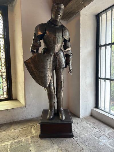 The image shows a full suit of ornate medieval armor displayed in an interior setting. The armor is silver with intricate etched designs. It stands upright on a dark wooden plinth. The background reveals stone walls with plaster trim and windows. The windows are adorned with decorative metal grilles and stained-glass accents. The location appears to be indoors, possibly a museum or historical building, but there are no people visible. The city of Tlalnepantla, Mexico is not apparent from the photograph.