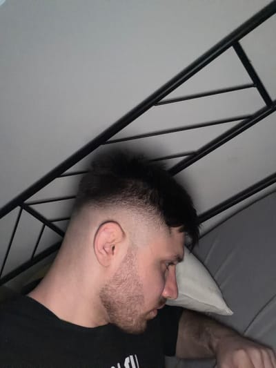 A man with a short, faded haircut and a beard lies on a pillow in a bed. He is wearing a black t-shirt. A black metal frame is in the background. The location is unknown and there is no information to indicate that it is in Pentre, United Kingdom.