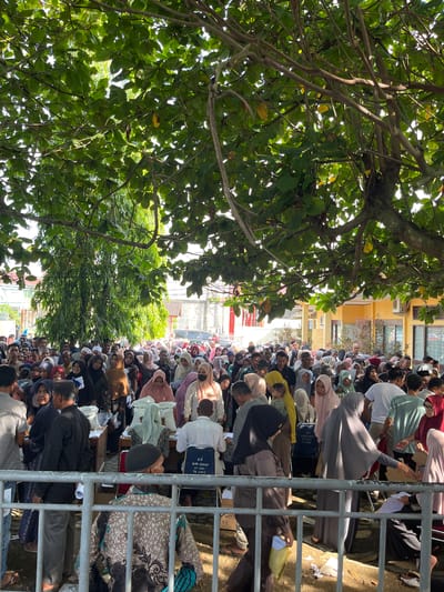A large crowd of people are gathered outdoors under a leafy tree, likely in Pulodolang, Indonesia. The scene appears to be some kind of distribution or event, with tables set up and bags of what might be supplies visible. Many individuals are wearing hijabs and traditional clothing. The atmosphere seems somewhat crowded and orderly, with people standing in lines and interacting with those at the tables. The environment is sunny, suggesting it's daytime, and the background includes buildings.