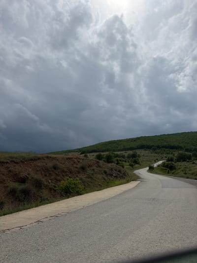 The image shows a paved road winding along a hillside near Drenkovo, Bulgaria. The road surface appears to be asphalt or concrete. The terrain on one side of the road is covered with vegetation, including trees and shrubs. On the opposite side, the sky is overcast with heavy gray clouds, suggesting an imminent weather event. The photograph appears to be taken from a low angle, close to the road surface. There are no visible people, animals, or vehicles.