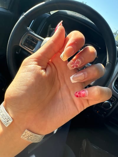 A human right hand, appearing to be female, is prominently featured within the interior of a vehicle. The hand displays a detailed manicure: the thumb and pinky nails feature a marbled pink and red pattern, while the index, middle, and ring fingers have clear/nude bases with white French tips. The middle finger is further adorned with a three-dimensional pink and white floral design. A silver-colored, open-cuff bracelet with embossed scrollwork is worn on the wrist. The hand is positioned over a black steering wheel, which has integrated media or cruise control buttons on its left spoke. Direct sunlight illuminates the hand, casting distinct shadows. Through the vehicle's windshield, a bright blue sky and indistinct green foliage are visible. The specific location within Rancho Cordova, United States, cannot be determined from the provided visual evidence.