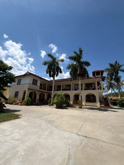 The image shows a two-story building with a light yellow facade and a red tile roof, situated in Hội Tín, Vietnam. The building has arched openings on the ground floor and balconies on the second floor, supported by pillars wrapped in decorative yellow and white fabric. Several tall palm trees are present on the grounds, with lush green foliage. The area in front of the building is paved with concrete, and there are some potted plants and small trees scattered around. The sky is a clear blue with scattered white clouds. The scene appears to be an outdoor common area, possibly a hotel or a public building, with no people visible. The overall environment suggests a warm, sunny climate typical of Vietnam.