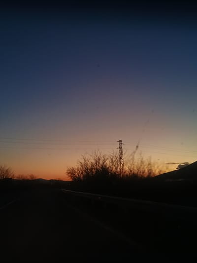 The image depicts a rural landscape near Zimnitsa, Bulgaria, captured during twilight. The sky transitions from a deep blue on the left to a warm orange near the horizon on the right. An electrical transmission tower stands in the middle ground, its wires stretching across the sky. A line of trees and bushes is silhouetted against the light, indicating the presence of vegetation. The foreground is dominated by a dark, undefined mass, obscuring details.